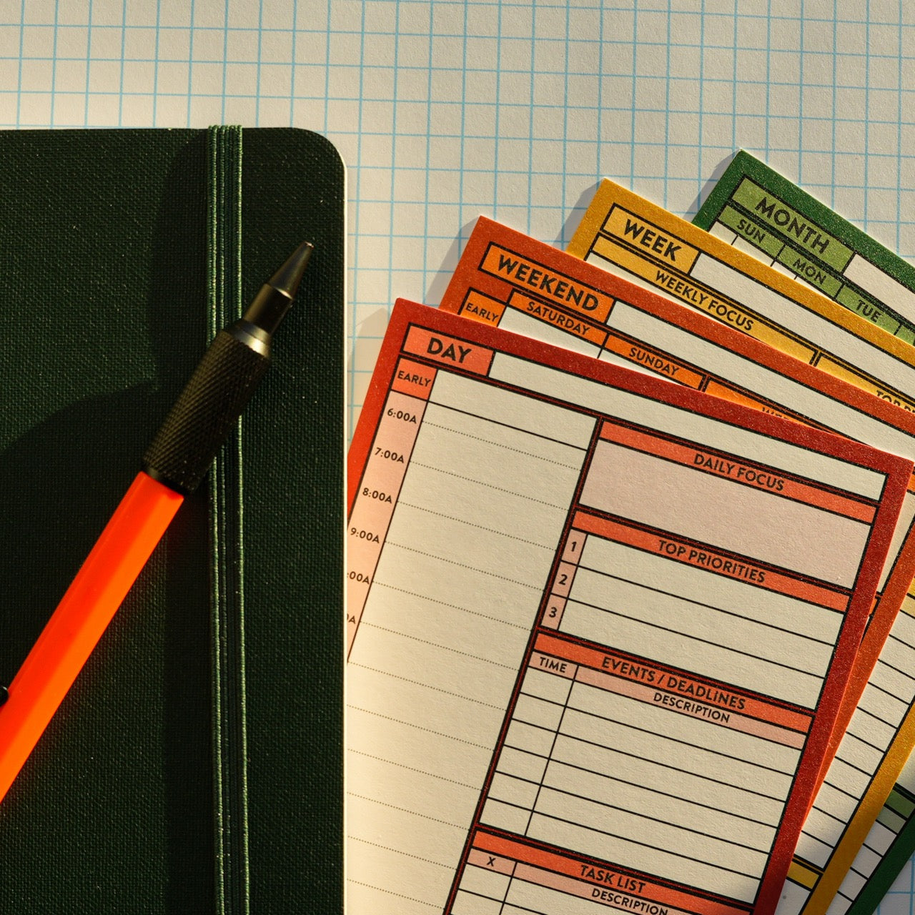 Day, Weekend, Week, and Month Better Made Plans planning cards laying next to a notebook with an elastic closure and a pencil on a graph paper background.