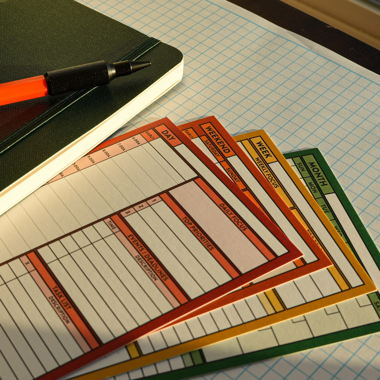 Day, Weekend, Week, and Month Better Made Plans planning cards laying next to a notebook with an elastic closure and a pencil on a graph paper background.