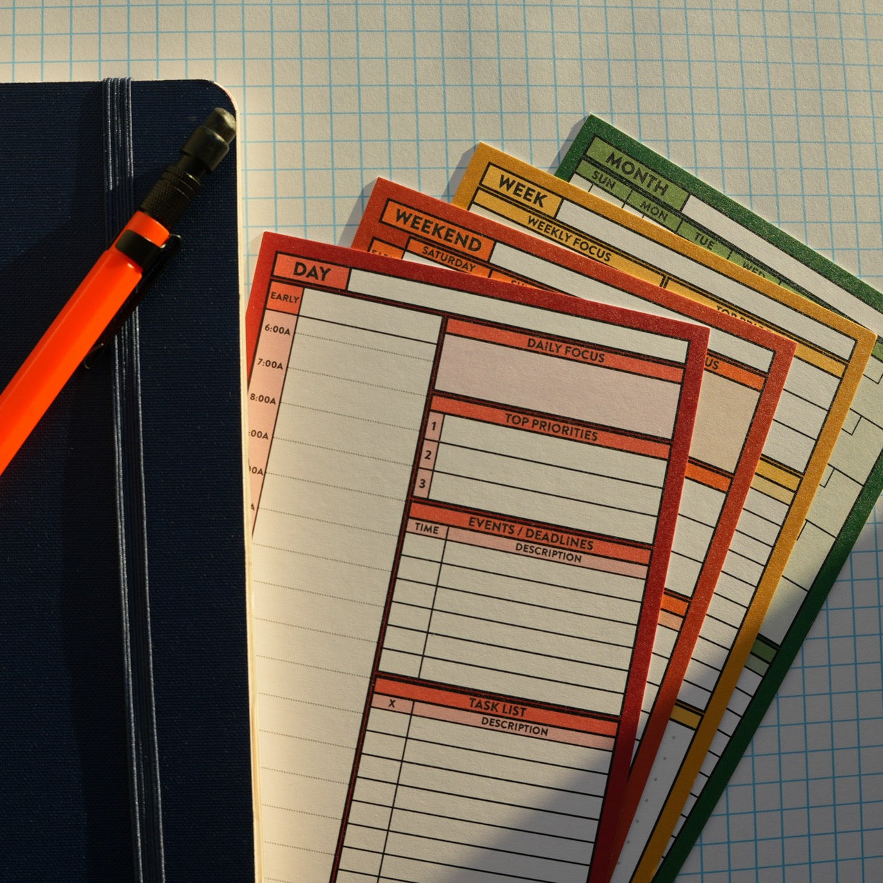 Day, Weekend, Week, and Month Better Made Plans planning cards laying next to a notebook with an elastic closure and a pencil on a graph paper background.