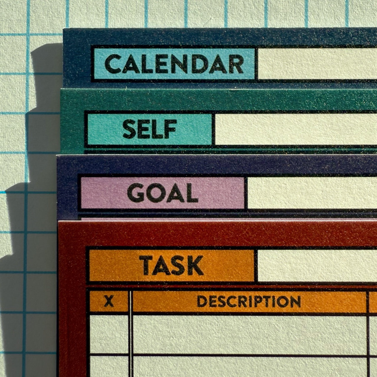 Picture of upper left corner labels of stacked Calendar, Self, Goal, and Task planning cards with a graph paper background.