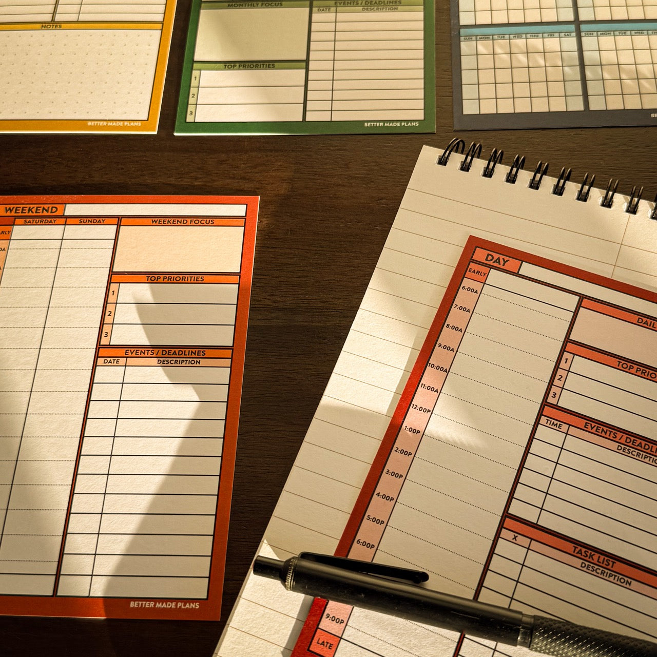 Various Better Made Plans planing cards laying on a wooden table with a notebook and pencil.