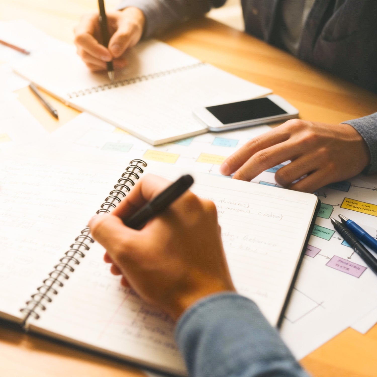 Realistic illustration of two individuals sitting at a table with notebooks, pens, and papers appearing to be collaborating or a meeting.